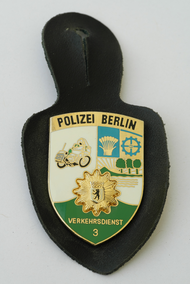 Ein Polizeistern mit der Aufschrift "Polizei Berlin", der ein blaues Schild mit einem weißen Stern in der Mitte zeigt, umgeben von einem weißen Rand, der auf einer Oberfläche liegt.
