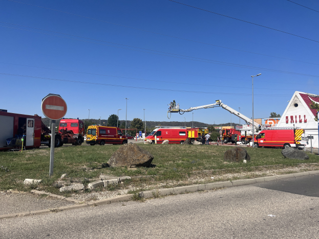 Eine Gruppe von Feuerwehrautos auf der Seite einer Stra├če neben einer Tankstelle nach einem Brand geparkt, mit umgebenem Gras, Steinen, einem Schild, einem Haus, Masten, einem Kran, Dr├Ąhten, B├Ąumen und einem bew├Âlktem Himmel.