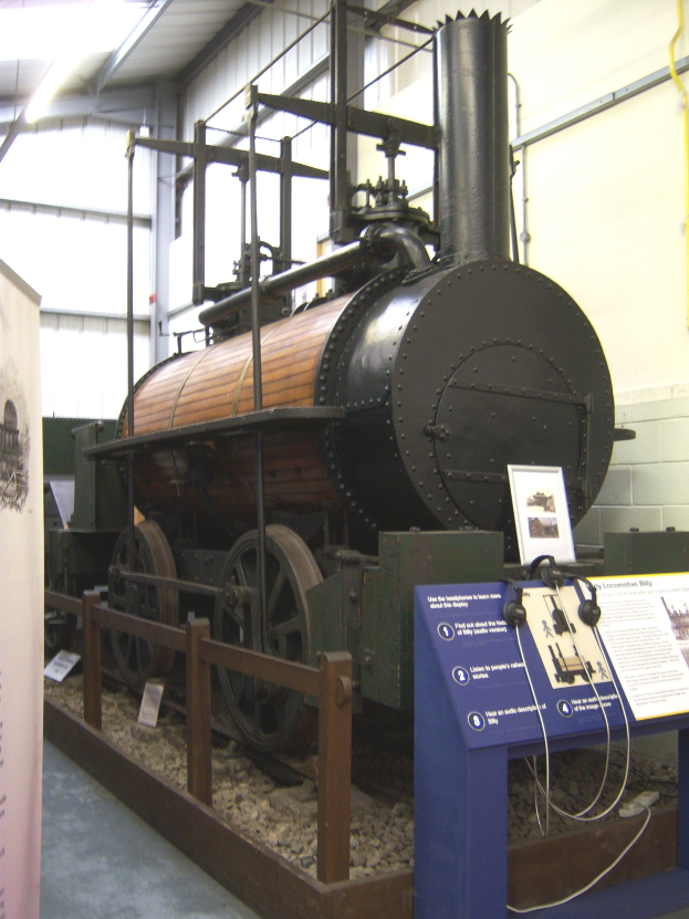 Alte Dampflokomotive in einem Museum mit Informationsschildern, einem Zaun, einer Fahne, Wandrohren und Deckenbeleuchtung.