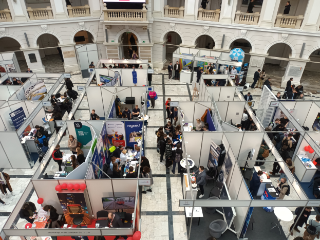 Ein voller Raum mit Ständen, Bannern, Tischen, Stühlen, Luftballons und einem Gebäude mit Bögen, Säulen, Geländern und einem Hintergrundschirm, was auf eine internationale Handelsmesse hindeutet.