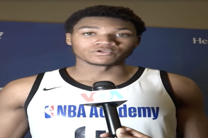 Ein Mann in einem weißen Tanktop hält ein Mikrofon und spricht mit der Medien bei der NBA All-Star Game 2019, mit einem Banner und einer Wand im Hintergrund.