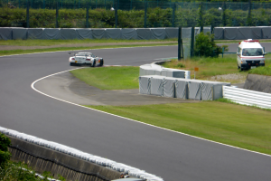 Ein Rennwagen fährt auf einer Rennstrecke mit einem Polizeiwagen im Hintergrund, umgeben von Vegetation und Absperrungen, mit Text unten.