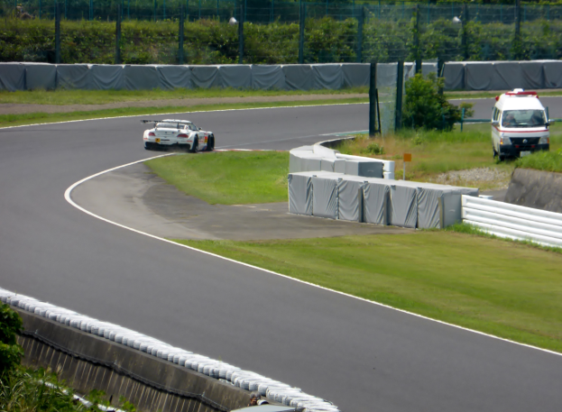 Ein Rennwagen fährt auf einer Rennstrecke mit einem Polizeiwagen im Hintergrund, umgeben von Vegetation und Absperrungen, mit Text unten.