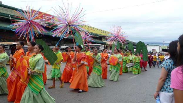Eine Gruppe von Menschen in farbenfrohen Kostümen marschiert während eines Karnevalsumzugs durch eine Straße, wobei eine Frau auf der rechten Seite eine Flasche hält, und Fahrzeuge, Gebäude, Bäume, Pfähle, Drähte und der Himmel im Hintergrund zu sehen sind.