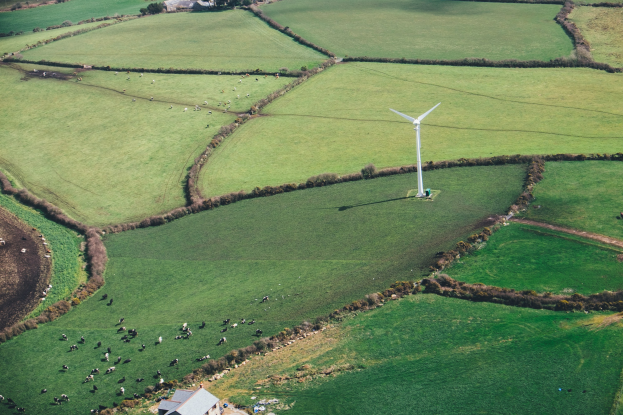 Luftaufnahme einer Windturbine in einer grünen Wiese mit Bäumen, Häusern und Tieren in Irland.
