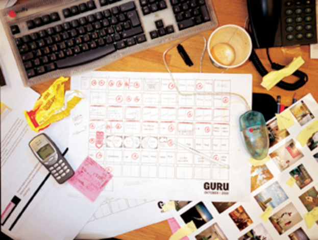 Ein chaotischer Schreibtisch mit Papieren, einem Computer, einem Telefon, Tassen und einem Handy.