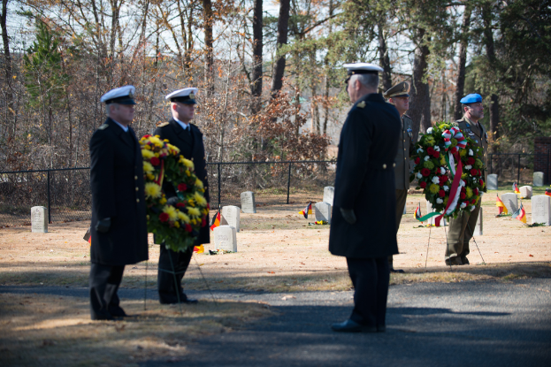 Zwei Beamte halten einen Kranz und stehen auf einem steinbedeckten Boden mit Flaggen, während ein weiterer Beamter vor ihnen steht und zwei weitere Beamte einen Kranz auf der rechten Seite halten, mit Bäumen im Hintergrund.