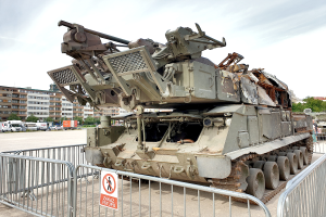 Ein gepanzertes Fahrzeug steht vor einem Metallzaun mit einem daran befestigten Schild. Im Hintergrund sind Fahrzeuge, Bäume, Gebäude und ein klarer blauer Himmel zu sehen.