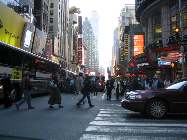 Eine belebte Stadtstraße mit Fahrzeugen, Fußgängern, die an einer Zebquerung überqueren, Gebäuden, Schildern, Verkehrsampeln und einem klaren blauen Himmel.