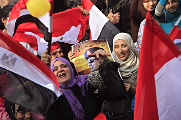 Eine Gruppe ägyptischer Demonstranten auf dem Tahrir-Platz schwenkt Flaggen und skandiert Parolen, während einige Schals tragen und einer ein Schild hält, während im Hintergrund ein Ballon zu sehen ist.
