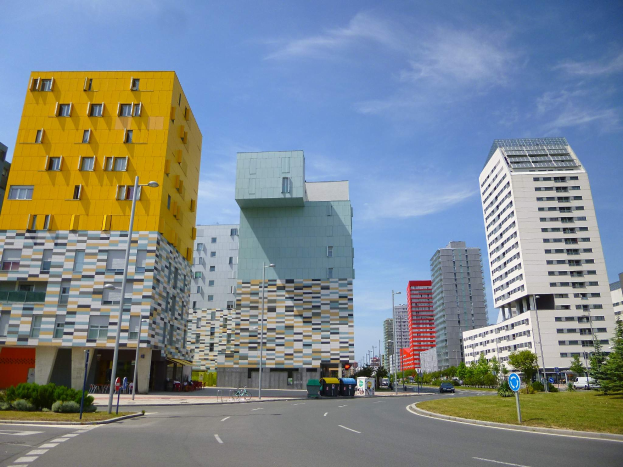 Eine Stadtstraße mit hohen, einzigartig gestalteten Universitätsgebäuden im Hintergrund, Straßenlaternen, Straßenlaternen, Kraftfahrzeugen, Informationsschildern, Büschen, Bäumen und einem bewölkten Himmel.