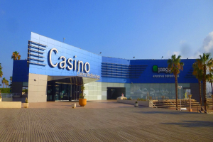 Ein Casino-Gebäude mit Palmen davor, umgeben von Topfpflanzen, einer Treppe, einem Geländer, einer Baumgruppe, einigen Pfählen und einem bewölkten Himmel.