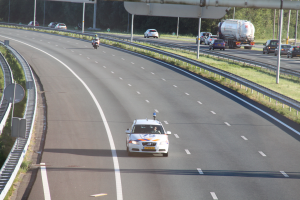 Ein Auto fährt auf einer Autobahn mit einem Polizeiwagen am Straßenrand, anderen Fahrzeugen, Leitplanken, Pfählen, Planken und Bäumen im Hintergrund.