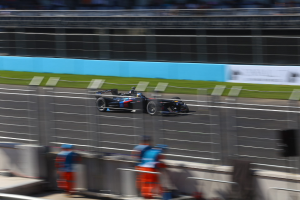 Ein fokussiertes Formel-Auto, das auf einer Rennstrecke fährt, mit unscharfen Zuschauern und grasbewachsenem Hintergrund, mit Texttafeln im Hintergrund.