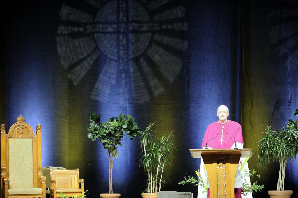 Ein Mann in einem rosa und weißen Kleid steht neben einem Podium in einem Auditorium, mit Pflanzen und zwei Stühlen daneben und einer bunten Wand im Hintergrund.