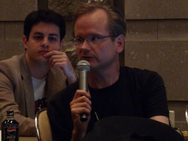 Zwei Personen sitzen auf Stühlen, eine trägt ein Jackett und die andere ein schwarzes T-Shirt und hält ein Mikrofon, links daneben eine Flasche mit schwarzer Kappe und einem Markennamen.