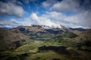 Panoramischer Blick von der Spitze eines Berges in Queenstown, Neuseeland, mit saftig grünem Gras, verstreuten Bäumen und einer gewundenen Straße in der Landschaft, unter einem Himmel voller weißer, flauschiger Wolken.