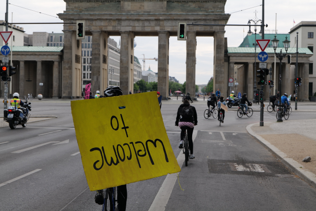 Eine Gruppe von Radfahrern fährt an der Brandenburgertor in Berlin vorbei, einer hält ein gelbes Schild, mit Laternenmasten, Verkehrsampeln, Gebäuden und Bäumen im Hintergrund bei klarem Himmel.