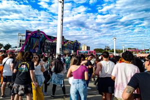 Eine Menschenmenge, einige mit Mützen und Goggles, steht vor einem Karneval mit Pfählen, Bäumen, Gebäuden und einem bewölkten Himmel im Hintergrund.