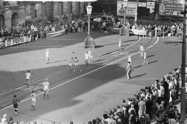 Schwarzes Bild eines Straßenfußballspiels mit Zuschauern, umgeben von Laternenmasten, Gebäuden und Bannern.