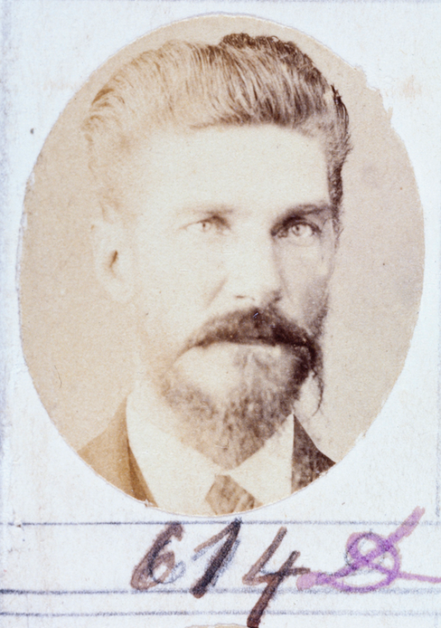 Ein altes Foto eines bärtigen Mannes mit Schnurrbart, der direkt in die Kamera schaut, mit leicht geöffneten Lippen, ordentlich gekämmtem Haar und einem gut gepflegten Bart.