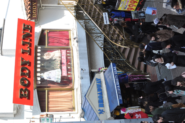 Eine Gruppe von Menschen, die auf einem Weg außerhalb gehen, mit einer Frau in einem schwarzen Kleid, die rechts einen weißen Stoff hält, einem Geschäft mit Schaufensterpuppen unten, zusätzlichen Schaufensterpuppen in der Nähe, einer Tafel mit eingraviertem Text links, einer Treppe in der Mitte und Tafeln mit gedrucktem Text oben.