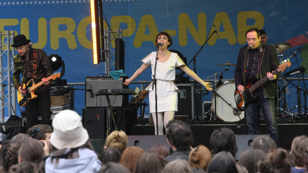 Eine Bühne mit einer Frau in einem weißen Kleid, die in der Mitte singt, zwei Männer mit Gitarren rechts und links und eine Menge unten sowie Lautsprecher im Hintergrund.