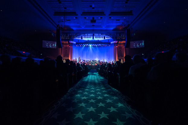 Ein großer Auditorium mit sitzenden Menschen, eine Bühne mit Musikern, die Instrumente spielen, und eine Decke mit Lichtern und Fernsehern, die einen Konzertsaal für die Nationale Convention 2019 suggerieren.