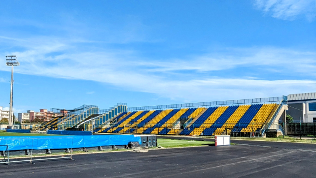 Ein Fußballfeld mit blauen und gelben Sitzplätzen, Geländern, Pfosten, Lampen, Bäumen, Gebäuden und einem bewölkten Himmel, mit einem Müllcontainer im Vordergrund.