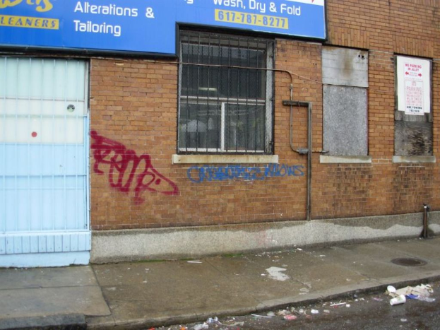 Eine Straßenszene mit einem Gehweg und einer Straße, die mit Müll übersät ist, einer Backsteinmauer, Fenstern, Rohren, einem Tor und einer Wandtafel oben.