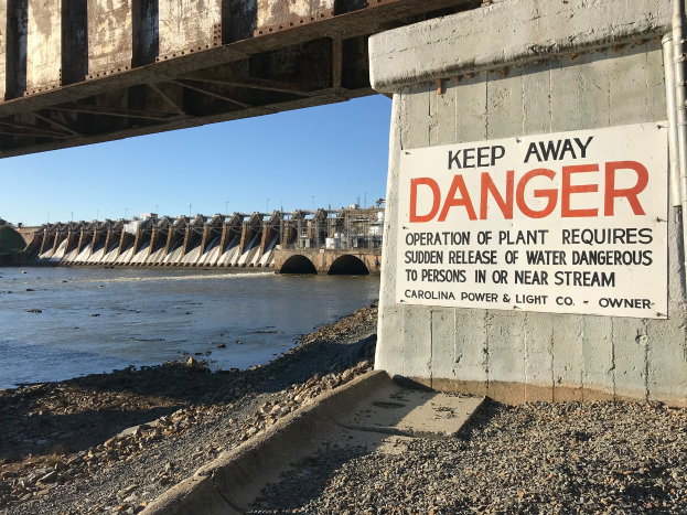 Eine Brücke mit einem "Vorsicht"-Schild, umgeben von Wasser, mit Gebäuden und einem klaren blauen Himmel im Hintergrund.