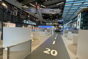 Ein großer Raum mit weißen Wänden, ein vom Himmel hängendes Flugzeug, ein Geländer auf der linken Seite, Gegenstände auf der rechten Seite, Texttafeln im Hintergrund und an Metallstangen an der Decke befestigte Lampen, wahrscheinlich am Düsseldorf Airport.