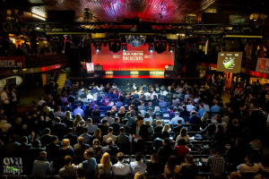 Eine große Menschenmenge, die auf Stühlen vor einer Bühne mit Bildschirmen und Bannern sitzt, die von Scheinwerfern beleuchtet wird und von Geländern umgeben ist, voller Erwartung auf den Beginn eines Online-Marketing-Rockstars-Events, mit einem Wasserzeichen in der linken unteren Ecke.