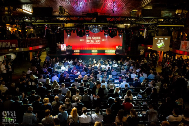 Eine große Menschenmenge, die auf Stühlen vor einer Bühne mit Bildschirmen und Bannern sitzt, die von Scheinwerfern beleuchtet wird und von Geländern umgeben ist, voller Erwartung auf den Beginn eines Online-Marketing-Rockstars-Events, mit einem Wasserzeichen in der linken unteren Ecke.