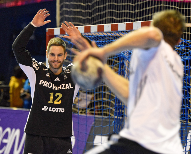Zwei Männer spielen Handball vor einem Tor mit Netz und Banner im Hintergrund und einigen Zuschauern in einem Stadion.