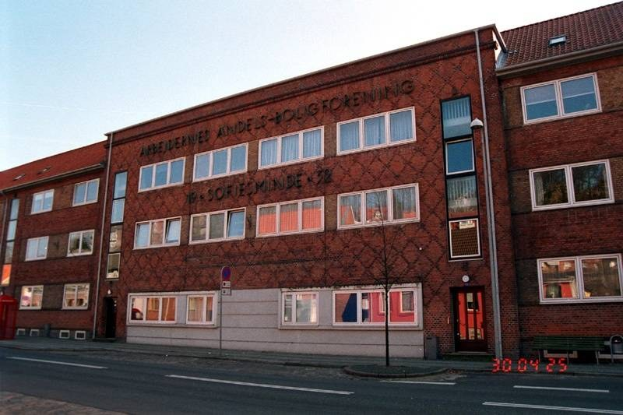 Großes Backsteingebäude mit vielen Fenstern, als Hauptquartier der deutschen Bundespolizei identifiziert, das sich auf einer Straße mit einem Gehweg, einem Baum, einer Bank, einem Laternenpfahl und einem roten Briefkasten befindet, vor einem Himmelhintergrund.