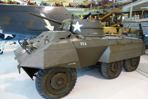 Ein gepanzertes Fahrzeug in einem Museum ausgestellt, umgeben von anderen Militärfahrzeugen, mit Menschen, Treppen, Geländern und Lichtern im Hintergrund.