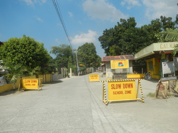 Gelbes "Schule"-Schild auf der linken Seite einer Straße mit parkenden Fahrzeugen auf der rechten Seite, Bäumen, Gebäuden, Strommasten, Drähten und Himmel im Hintergrund.