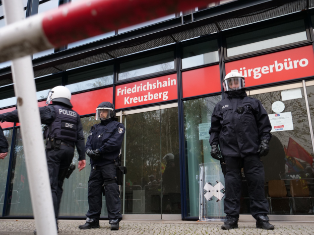 Polizeibeamte in Uniform stehen vor einem Gebäude mit Glaswänden und -türen, tragen Helme und Handschuhe, mit einem Pfahl links und Schildern, die "Friedrichshain Kreuzberg" im Hintergrund lesen.