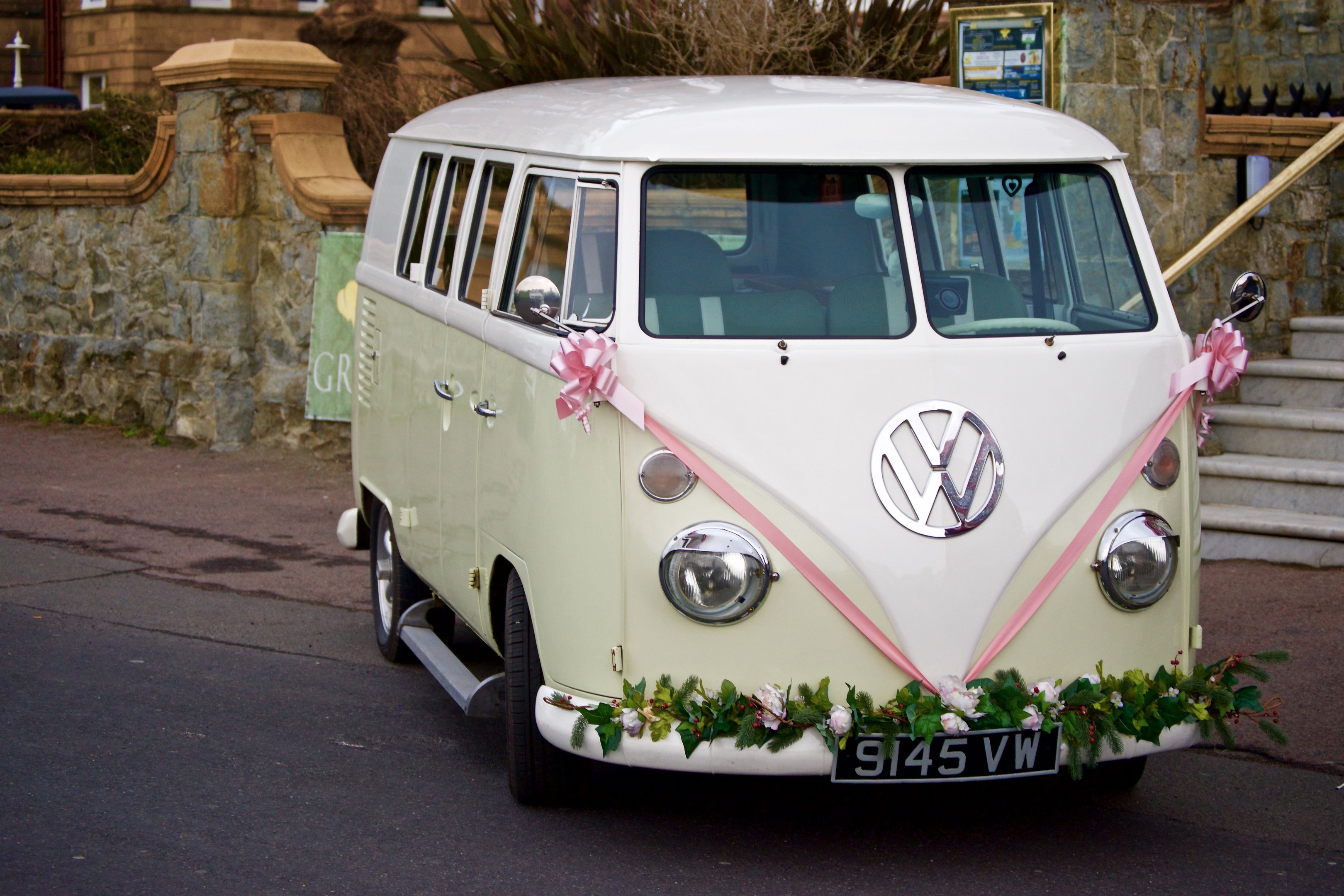 Ein weißer und pinker VW Camper Van mit floralen Verzierungen, der vor einer Steinmauer geparkt ist, neben einer Treppe und einem Gebäude im Hintergrund.