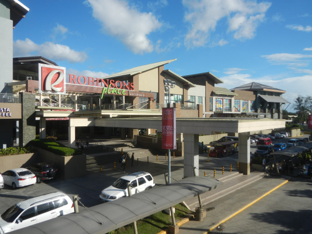 Ein belebter Einkaufszentrum mit Gebäuden, Schaufenstern, Fahrzeugen, Fußgängern, Straßenmöbeln, Grünflächen und einem bewölkten Himmel.