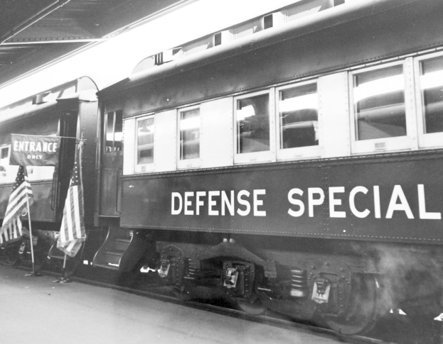Schwarz-weißes Foto eines Zuges mit der Aufschrift "Defense Special", der auf den Gleisen geparkt ist, mit zwei Fahnen links und einer Decke oben.