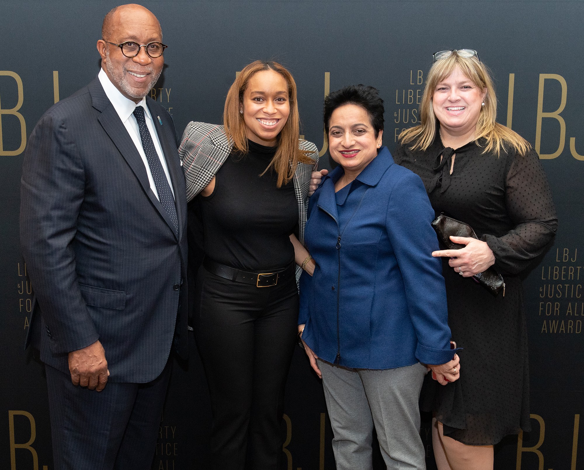 Eine Gruppe von vier Menschen auf einem roten Teppich, die lächeln und für ein Foto posieren, mit einer Tafel im Hintergrund, auf der "Liberty Justice for All Awards" steht.