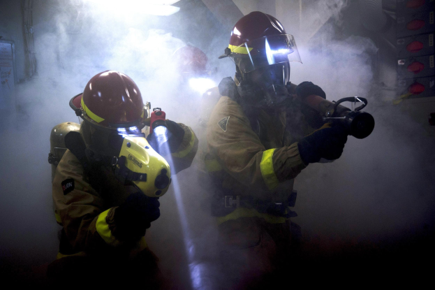 Zwei Feuerwehrleute in Schutzausrüstung sprühen Wasser auf eine rauchgefüllte Wand, mit verschiedenen Gegenständen rechts und einer Tafel mit Text links.
