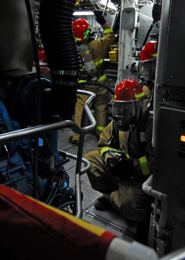 Feuerwehrleute in Helmen und Schutzausrüstung arbeiten an einem Feuerlöschwagen mit Rohren und Gegenständen im Hintergrund, unter einem Licht an der Spitze.