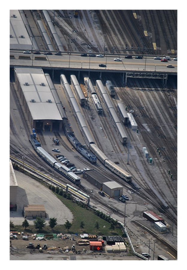 Eine Brücke mit einigen Fahrzeugen darauf, darunter Eisenbahnschienen mit einigen Zügen und eine Landschaft mit Gebäuden, Bäumen, Pfählen und verschiedenen Gegenständen und Fahrzeugen auf dem Boden.
