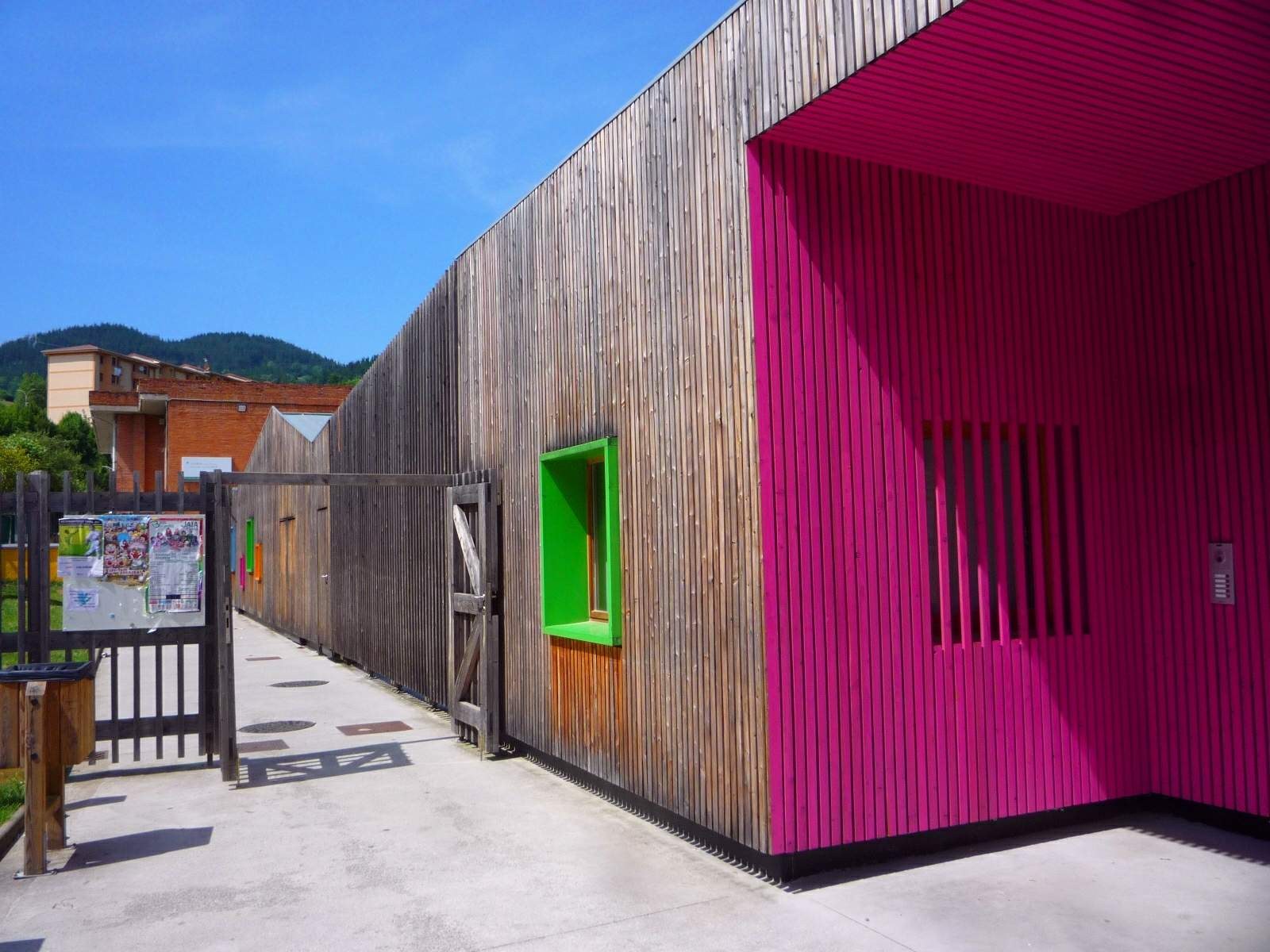 Ein pinkes Gebäude mit einer grünen Tür und Fenstern, einem hölzernen Zaun, einer Tafel mit Papieren, einem Müllcontainer, Gras, Bäumen, fernen Gebäuden, Hügeln und einem bewölkten Himmel, Teil eines neu umgebauten Schulspielplatzes in Spanien.