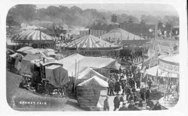 Ein altes Schwarz-Weiß-Foto eines lebhaften Karnevals mit Zelten, Menschen, Wagen, Pfählen, Bäumen und Gebäuden im Hintergrund, beschriftet mit "Barnet Fair, London, England".