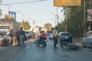 Eine Gruppe von Menschen steht um ein verunglütztes Motorrad auf der Seite einer Straße mit mehreren Fahrzeugen, darunter ein Lastwagen, und einem Hintergrund aus Bäumen, Masten, Lichtern und Schildern unter dem Himmel.