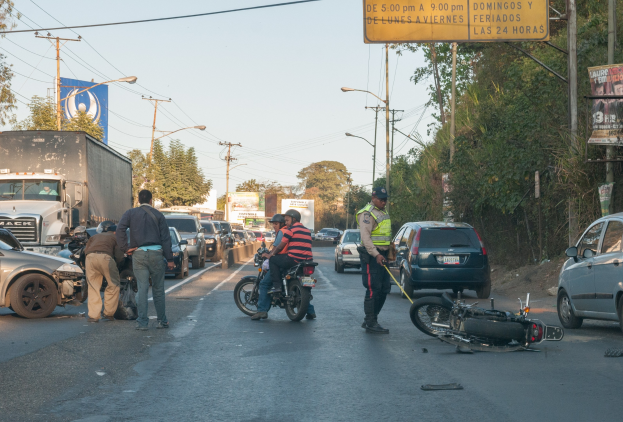 Eine Gruppe von Menschen steht um ein verunglütztes Motorrad auf der Seite einer Straße mit mehreren Fahrzeugen, darunter ein Lastwagen, und einem Hintergrund aus Bäumen, Masten, Lichtern und Schildern unter dem Himmel.